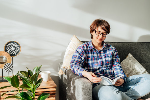 戴眼镜的女士坐在沙发上放松，在家里舒适的现代室内客厅里阅读一本书，绿色植物。线下活动的生活方式。阅读的爱好。休闲和娱乐。图片下载