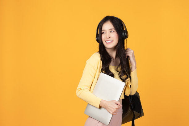 肖像快乐休闲的亚洲女生与背包，耳机和笔记本电脑孤立的黄色背景。回归学校和学习理念。图片下载