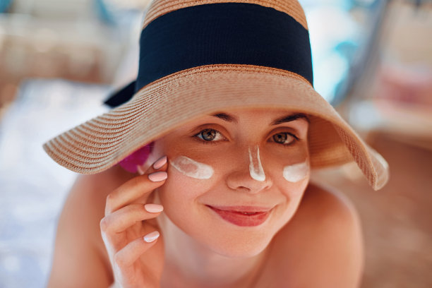 脸上涂着防晒霜的年轻女子拿着防晒霜瓶在海滩上。女性戴着帽子在皮肤上涂抹保湿乳液。图片下载