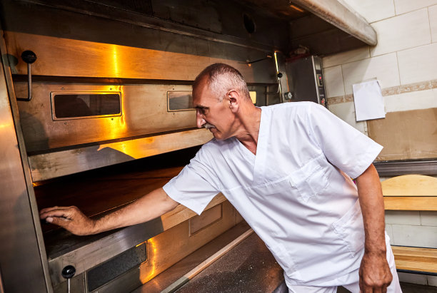 专业的面包师穿着统一的服装，在面包房的工业烤箱中放入烤生面团的面板图片下载