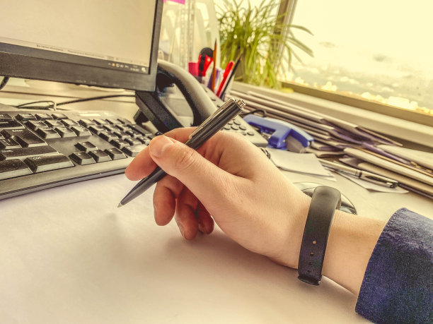 这位上班族手里拿着一支带杆的黑色金属笔。创建手写笔记。记事本放在键盘旁边图片下载