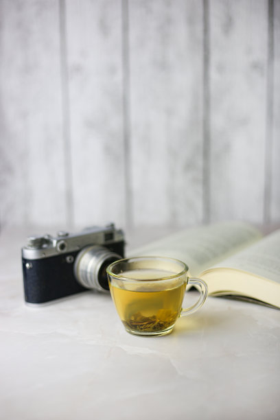 在一个透明的杯子里冲泡的花草绿茶，旁边是一本打开的书和一个光线背景上的老式相机图片下载