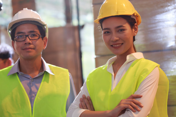 年轻迷人的亚洲女性戴着黄色的安全帽和安全背心与同事站在仓库，物流行业的概念。图片下载