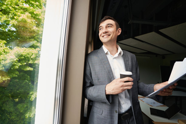 满意的男性办公室职员端着咖啡望向窗外图片下载