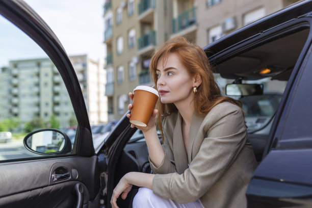 一名成年女子开着车门坐在车里，一边沉思一边用旅行杯喝着咖啡。图片下载