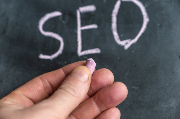 成年男子手里拿着一支彩色粉笔。手写字SEO在黑色黑板上。图片下载
