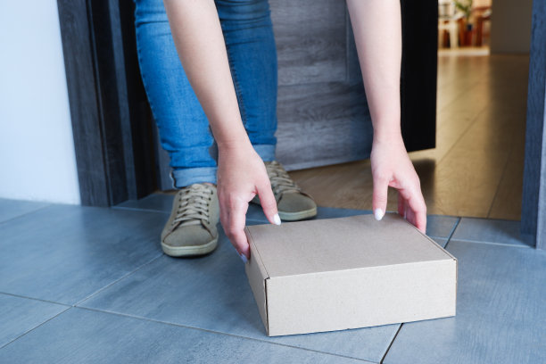 包裹送到门口。一个穿着蓝色牛仔裤的女人从门口拿起一个盒子，里面装着快递公司送来的货物。图片下载