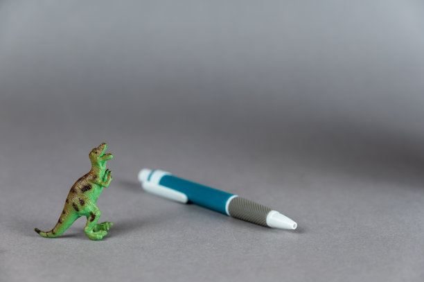 一只恐龙和一支圆珠笔在灰色背景下的微型模型。用后腿站立的食肉动物的绿色小雕像。White-gray-blue塑料笔。有选择性的重点。图片下载