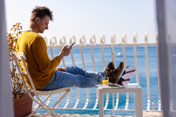 在酒店阳台上，一名男子坐在笔记本电脑旁用手机发短信图片下载