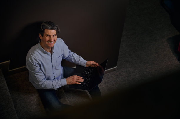 俯视一个英俊成熟的商人，企业家，文案在笔记本电脑上工作，坐在一个家庭内部的楼梯上，微笑着看着镜头露出牙齿的微笑图片下载