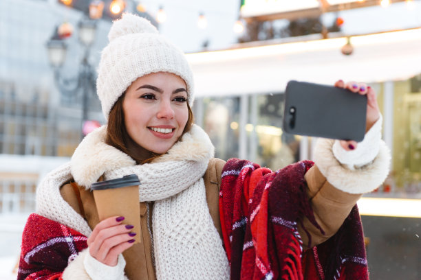 戴着帽子和围巾的年轻女子在冬天的雪地里喝着咖啡走在户外。图片下载