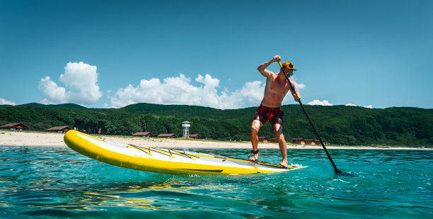 晒得黝黑的男人站在黄色的冲浪板上划桨。在碧绿的海水中，沙滩上进行着积极的运动图片下载