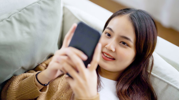 快乐的年轻亚洲女性放松在舒适的沙发在家里用智能手机发短信，微笑的女孩使用手机，浏览无线网络小工具，在家网上购物图片下载