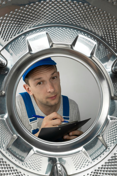 身穿制服的男修理工手里拿着一块平板，正在看洗衣机的滚筒，照片来自里面。垂直图图片下载