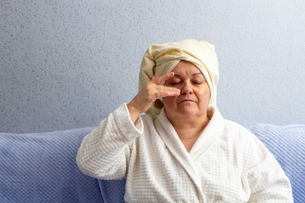 老妇人头上裹着毛巾，穿着浴袍，用手做面部按摩。贴片贴在脸上。有选择性的重点。图片为与年龄相关的面部护理的文章。图片下载