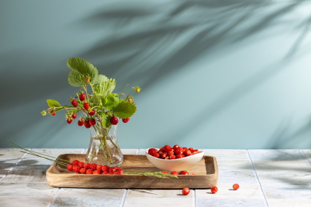 一束野生草莓和树叶在玻璃花瓶和许多浆果在木托盘。夏天静物与野生草莓在蓝色的背景。副本的空间。图片下载