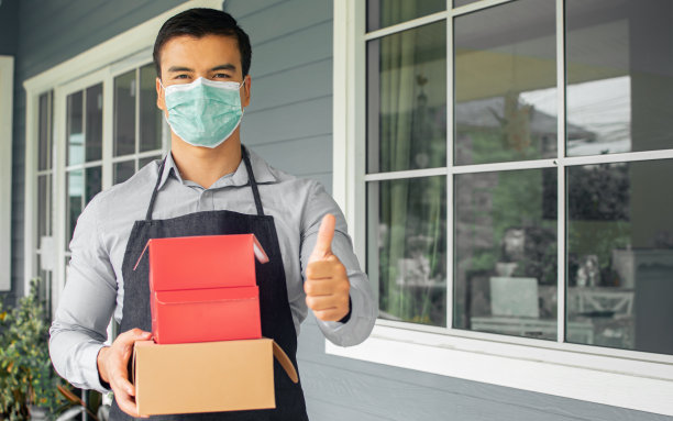 白人男性咖啡店和面包店老板，企业家戴着口罩保护病毒，拿着送货的盒子，站在商店外面。新常态，社会距离概念。图片下载