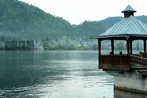 混凝土地基上的木制凉亭悬挂在一个大湖泊的水面上，另一边是针叶林中的一个小村庄。图片下载