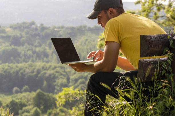 年轻人在网上工作时使用笔记本电脑，笔记本电脑在大自然的山林背景下户外。在笔记本电脑黑屏上复制设计空间或文本图片下载