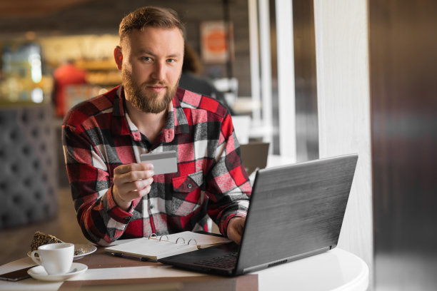 惊人的胡子年轻人的肖像坐在咖啡馆使用笔记本电脑和持有借记卡。微笑，用信用卡网购。图片下载