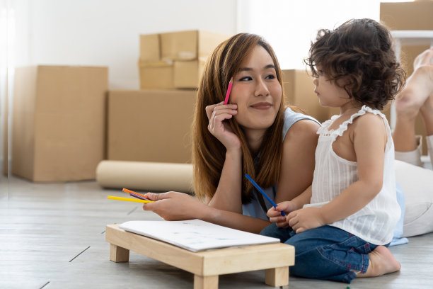 年轻的亚洲母亲在教她的小女儿在纸上画彩色铅笔坐在许多纸板盒在家里。孩子通过画画来学习。教育的概念图片下载