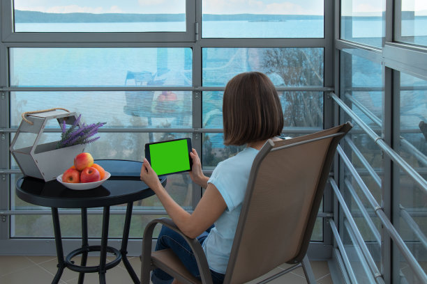 白人女性坐在扶手椅上，使用绿色屏幕的平板电脑。从后面看。舒适的地方放松与全景窗户图片下载