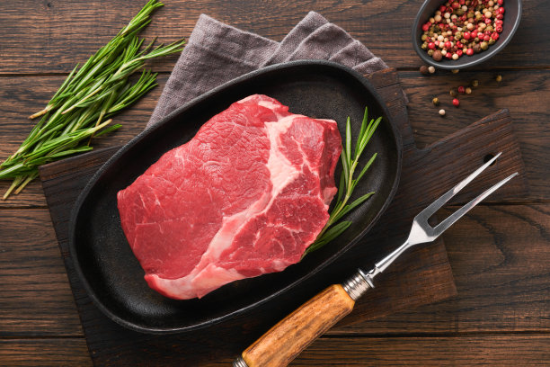 牛排。新鲜的生牛排肋眼牛排，叉迷迭香，盐和胡椒粉放在烤盘上，黑色旧的木制桌子背景。商店广告的想法。前视图。设计理念的模型。图片下载