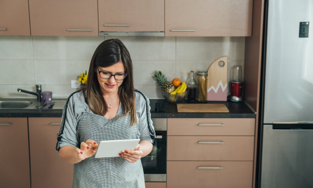 穿着围裙的微笑女人一边在现代厨房里做饭，一边在电子平板电脑上阅读菜谱图片下载