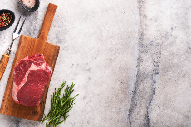 牛排。新鲜生牛肋眼牛排，叉迷迭香，盐和胡椒粉放在老木架子上，灰色的石头背景。前视图。设计理念的模型。图片下载