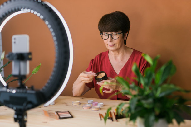 中年女性美妆博主或网红在家里的房间里用化妆产品录制视频。使用环形灯和智能手机图片下载