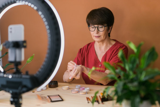 中年女性美妆博主或网红在家里的房间里用化妆产品录制视频。使用环形灯和智能手机图片下载