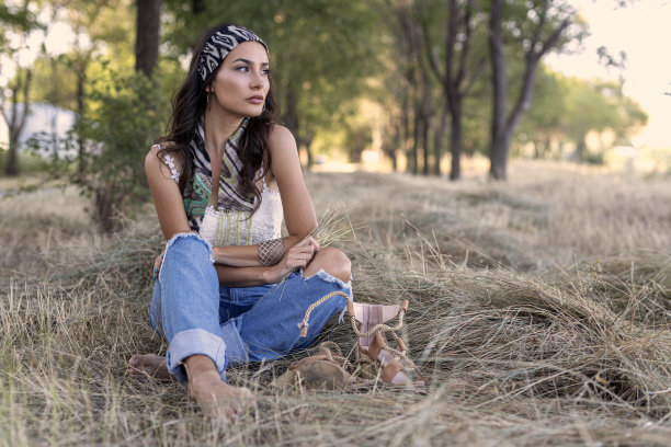 乡村时装女人，头上戴围巾，穿着牛仔裤，坐在田野里的稻草上图片下载