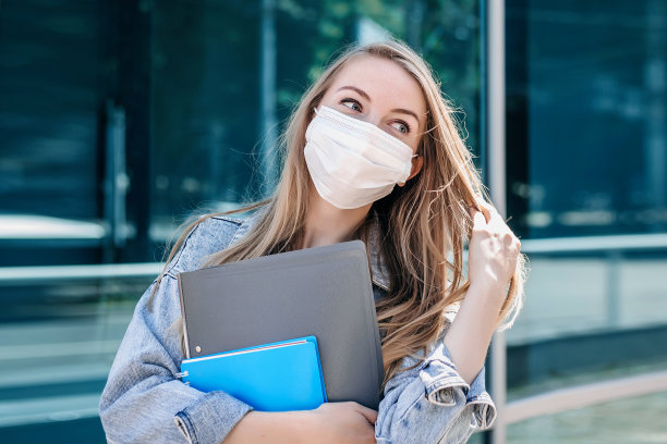 戴着防护口罩的年轻漂亮金发女孩站在一座玻璃幕墙的现代建筑的背景下，远程工作，covid-19，隔离图片下载