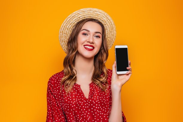 一名年轻女子穿着红色的夏季连衣裙，帽子拿着手机，向镜头展示它，黑色的空屏幕，模型，设计空间，在线订单，购物概念，复制空间，橙色背景图片下载