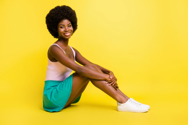 年轻快乐开朗微笑可爱的非洲裔女孩坐地板穿绿色裙子孤立在黄色背景照片图片下载