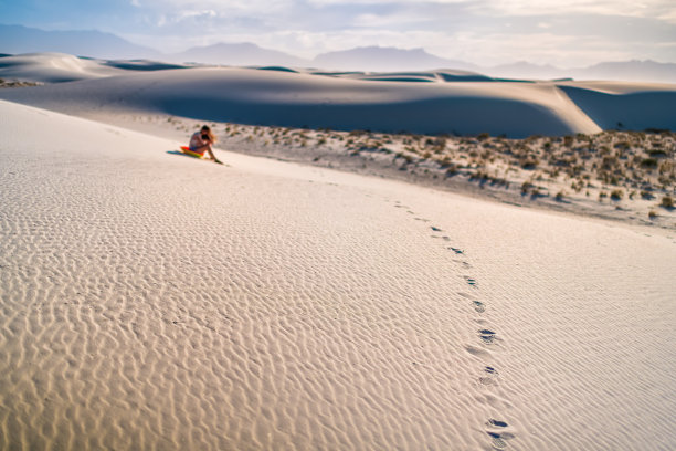 在新墨西哥州著名的白沙沙丘国家纪念碑上的年轻女子坐在圆盘雪橇在日落期间滑下山图片下载