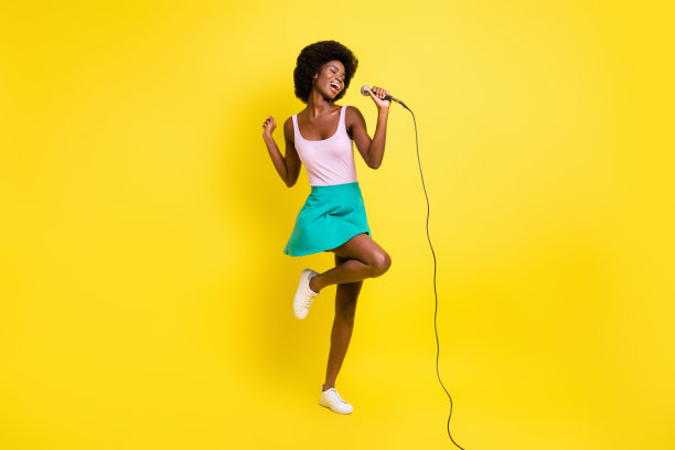 全身照片的深色皮肤有吸引力的女孩穿粉红色的背心举行唱歌麦克风隔离在黄色的背景图片下载
