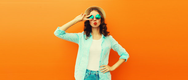 美丽的年轻女子的肖像，她的嘴唇发送甜蜜的空气吻摆着夏天的草帽在橙色的背景图片下载