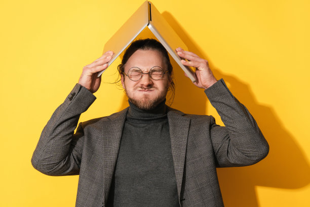 一名男子在黄色背景上做鬼脸，把笔记本电脑举过头顶图片下载