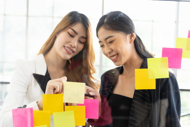 一群亚洲商界女性在办公室开会，并在玻璃墙上贴上便签。商业亚洲女性使用便利贴头脑风暴和分享想法在玻璃墙。图片下载