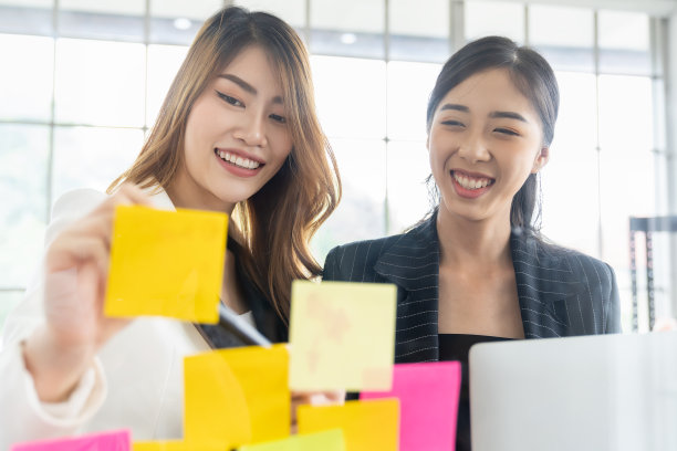 一群亚洲商界女性在办公室开会，并在玻璃墙上贴上便签。商业亚洲女性使用便利贴头脑风暴和分享想法在玻璃墙。图片下载