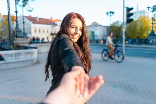 快乐的黑发女孩脸上带着微笑牵着男友的手在一个温暖的春天，夏天的傍晚日落的街道上。跟我来的概念。有选择性的重点。图片下载