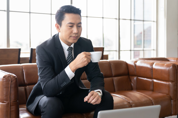 亚洲商人坐在沙发上喝咖啡，用笔记本电脑在办公室工作。图片下载