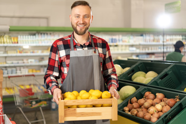 在超市拿着柠檬微笑的白人职员看着镜头，在杂货店的果蔬柜台购物，穿着围裙的男人拿着一箱柠檬站在超市购物图片下载