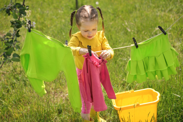 在一个阳光温暖的夏天，穿着鲜艳衣服的可爱女孩笑着把粉红色的t恤挂在晾衣绳上晾干。妈妈的小帮手。做家务图片下载