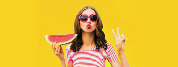夏天的肖像美丽的年轻女子吹她的嘴唇与红色的口红发送空中吻西瓜片戴着心形墨镜在黄色的背景图片下载