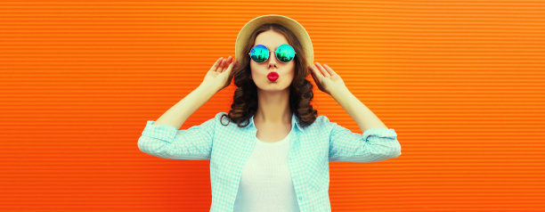 美丽的年轻女子的肖像，她的嘴唇发送甜蜜的空气吻摆戴夏天的草帽在橙色的背景，空白的拷贝空间的广告文字图片下载