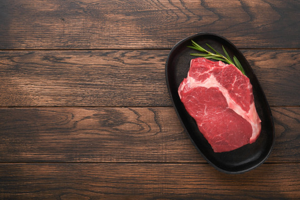 牛排。新鲜的生牛排肋眼牛排，叉迷迭香，盐和胡椒粉放在烤盘上，黑色旧的木制桌子背景。商店广告的想法。前视图。设计理念的模型。图片下载
