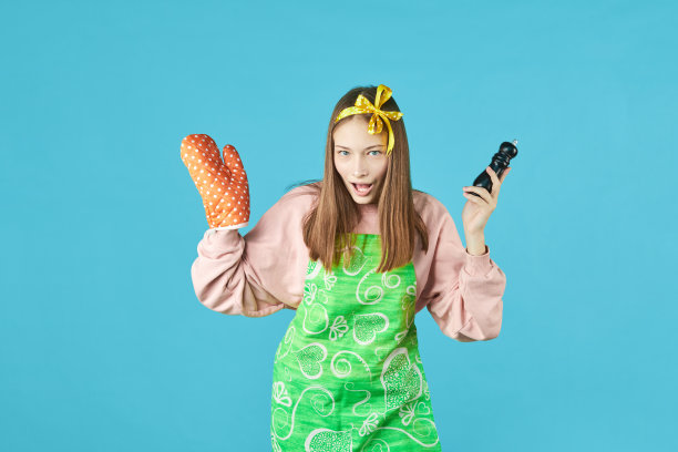 穿着厨房衣服的快乐女孩在蓝色的背景上拿着木勺和锅架。穿围裙的年轻女子是一个有厨房用品的家庭主妇。图片下载