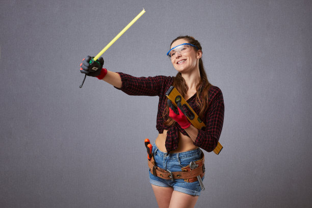 快乐的长发工人戴着防护眼镜。性感女职工手持卷尺。维修，施工和建筑概念-女孩与工作工具在灰色背景。图片下载